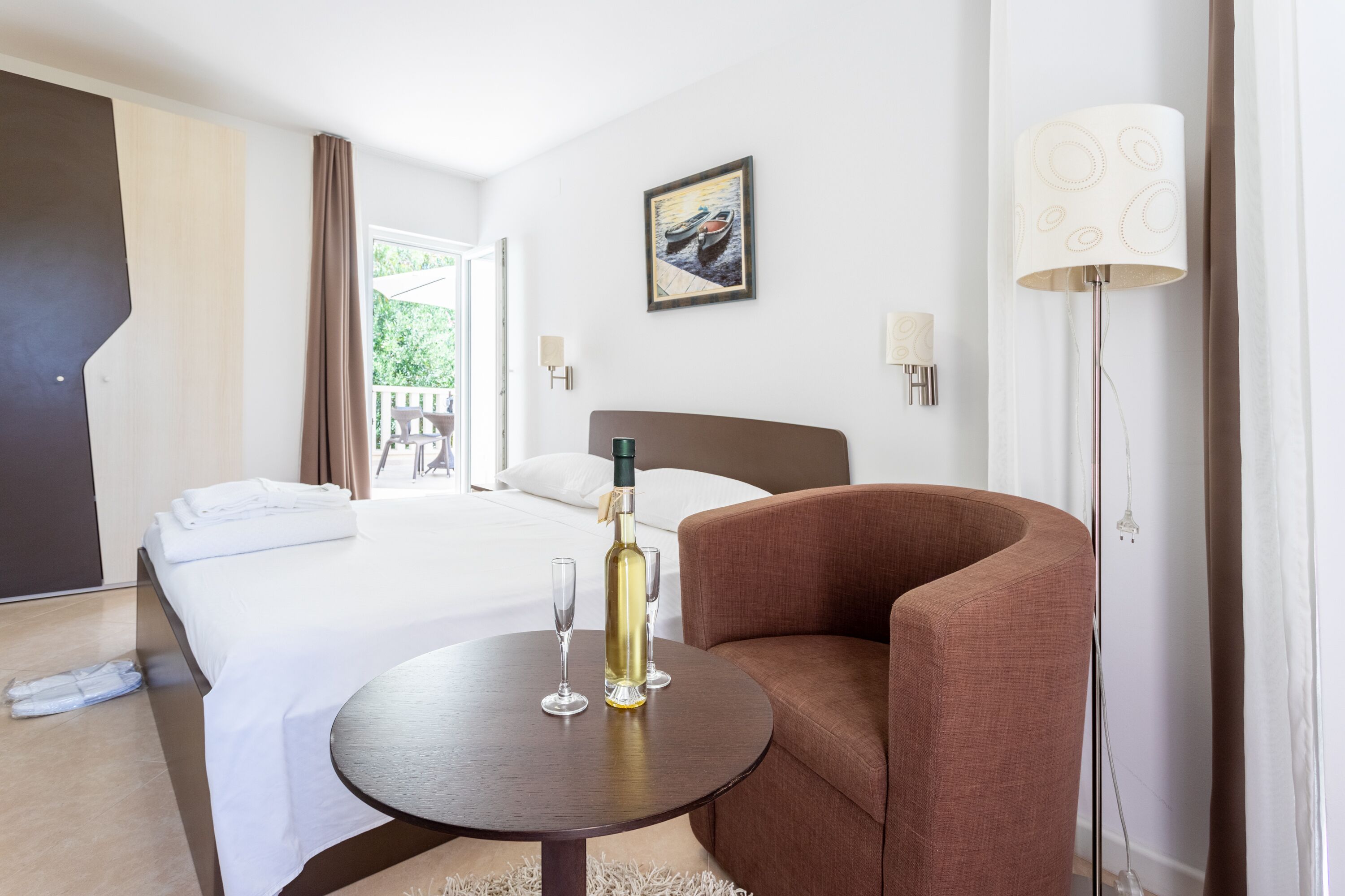 A hotel room with a bed, a round table with a bottle and glasses, a brown armchair, a painting on the wall, and a lamp.