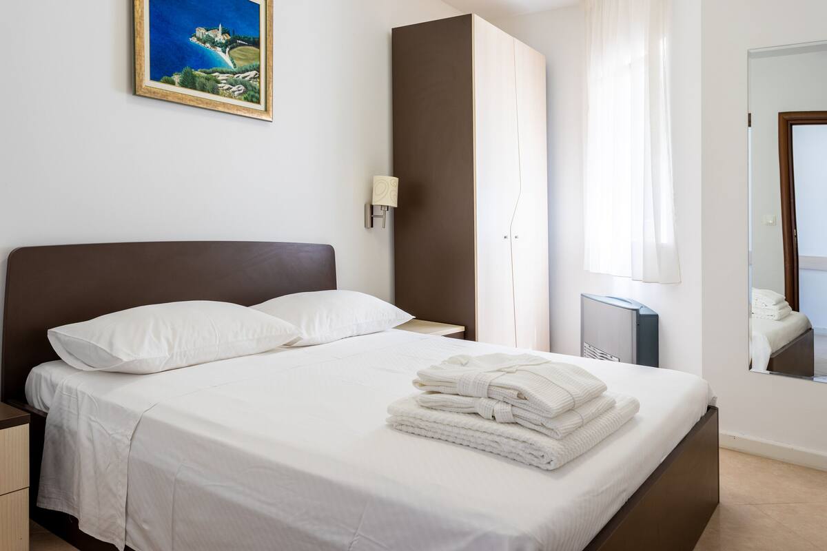 A neatly made bed with white linens, a nightstand with a lamp, a wardrobe, and a painting on the wall.