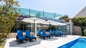 Un espace aménagé au bord de la piscine, avec des chaises longues et des parasols, une structure entièrement vitrée et une piscine à l’eau d’un bleu limpide.