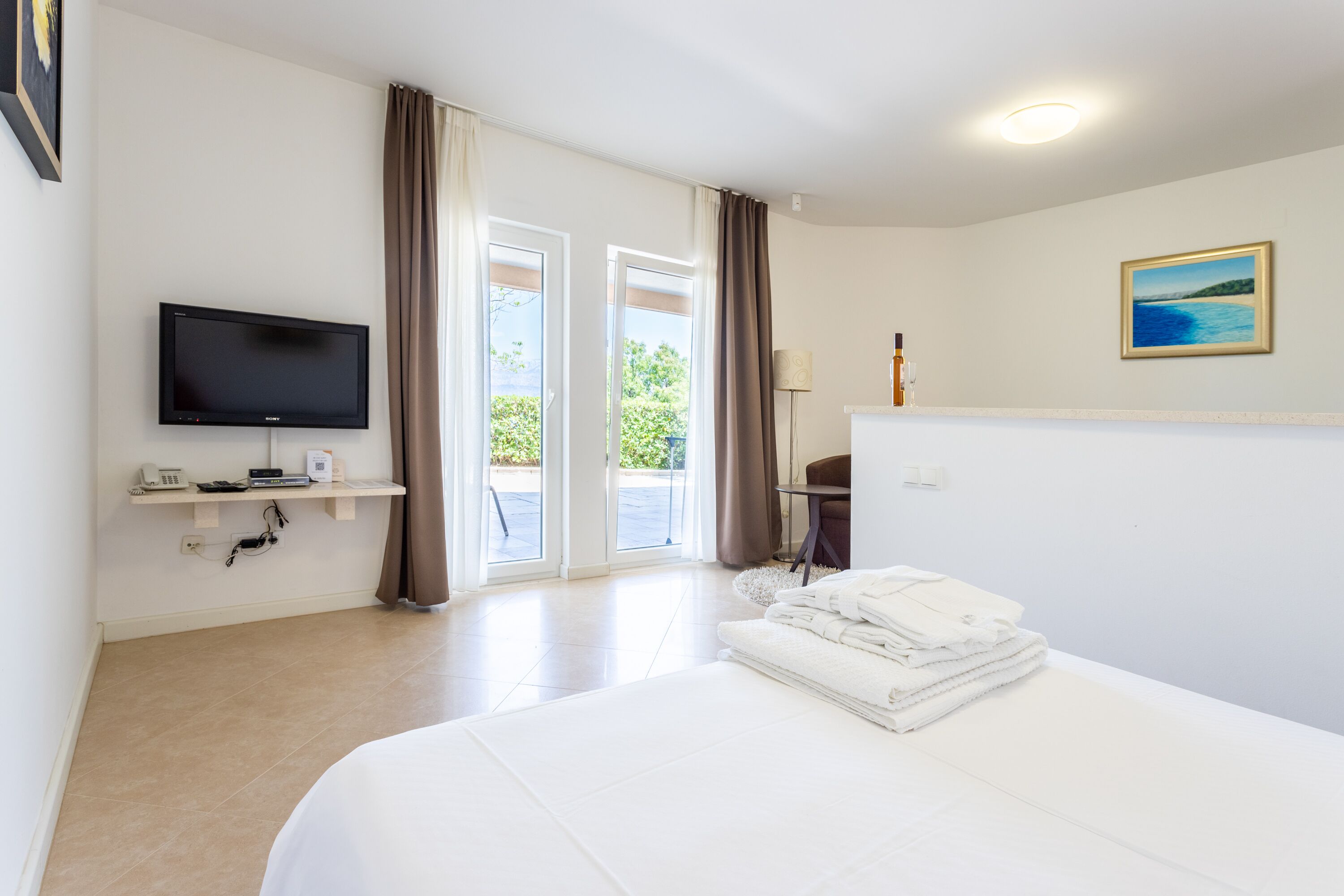 A hotel room with a bed, a TV, a desk with a lamp, a chair, and a view of the outdoors.
