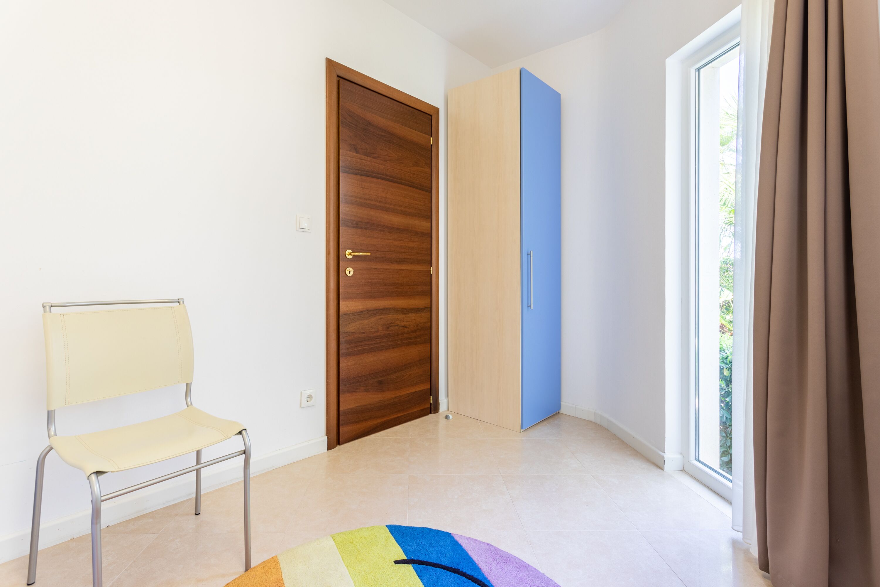 A room with a wooden door, a blue wardrobe, a white chair, and a large window with curtains.