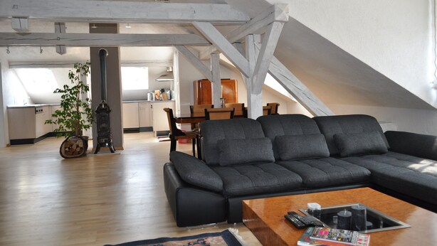Un appartement moderne de style « loft », avec des poutres en bois apparentes, un canapé en cuir noir, une table basse en bois et une cuisine en arrière-plan.