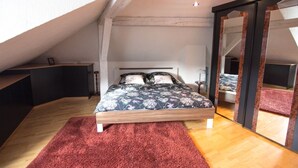 Une chambre avec un plafond en pente, un lit recouvert d’une literie à motifs floraux, un tapis rouge et un grand miroir.