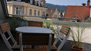 Un balcon avec une table et des chaises en bois, des plantes en pot et une vue sur des immeubles et une colline.
