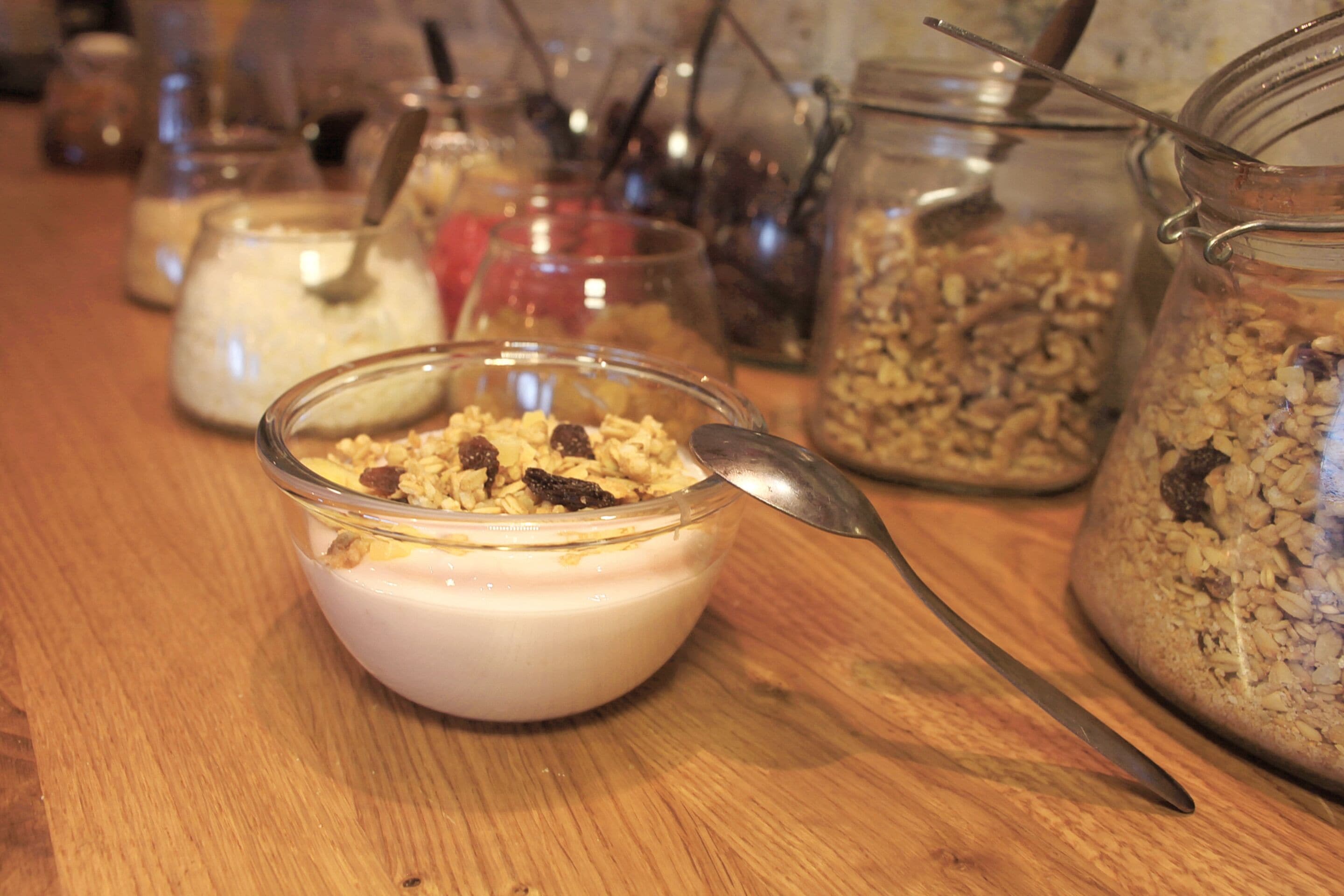 A wooden table with various food items, including a bowl of yogurt with granola and a spoon.