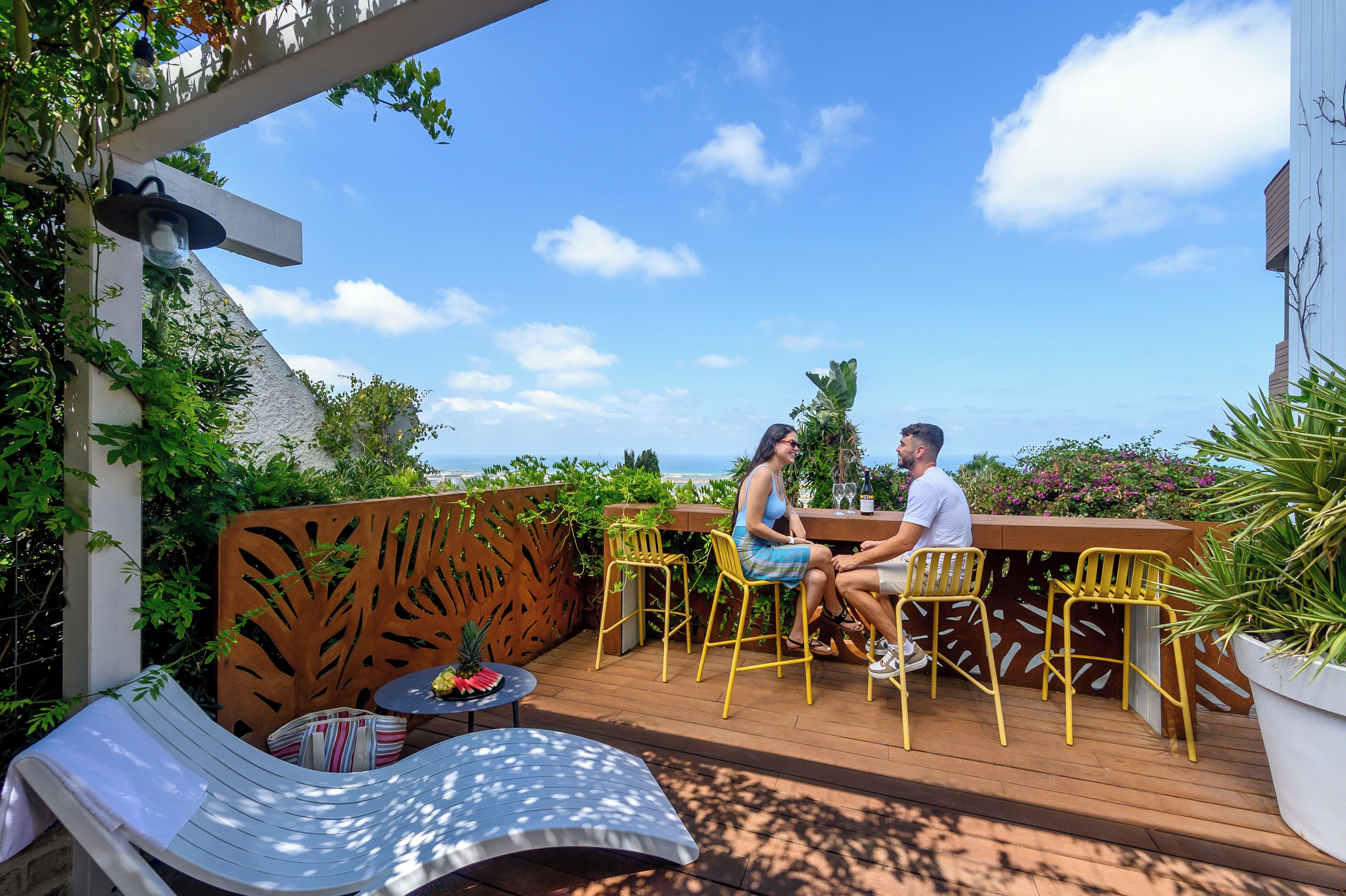 A couple sitting at a wooden deck with yellow chairs, a white table, and a striped cushion.