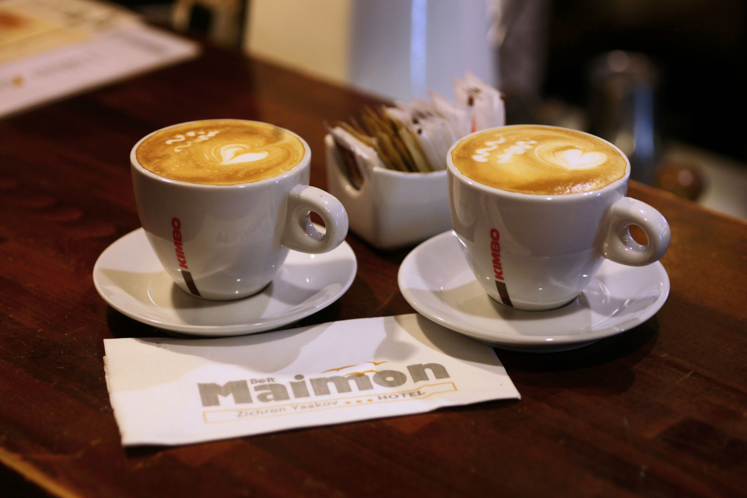 Two cups of coffee with heart-shaped latte art on a wooden table, accompanied by a napkin with the hotel name.