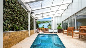 Una piscina con plataforma de baldosas, un porche cubierto y un muro de ladrillo con plantas trepadoras.