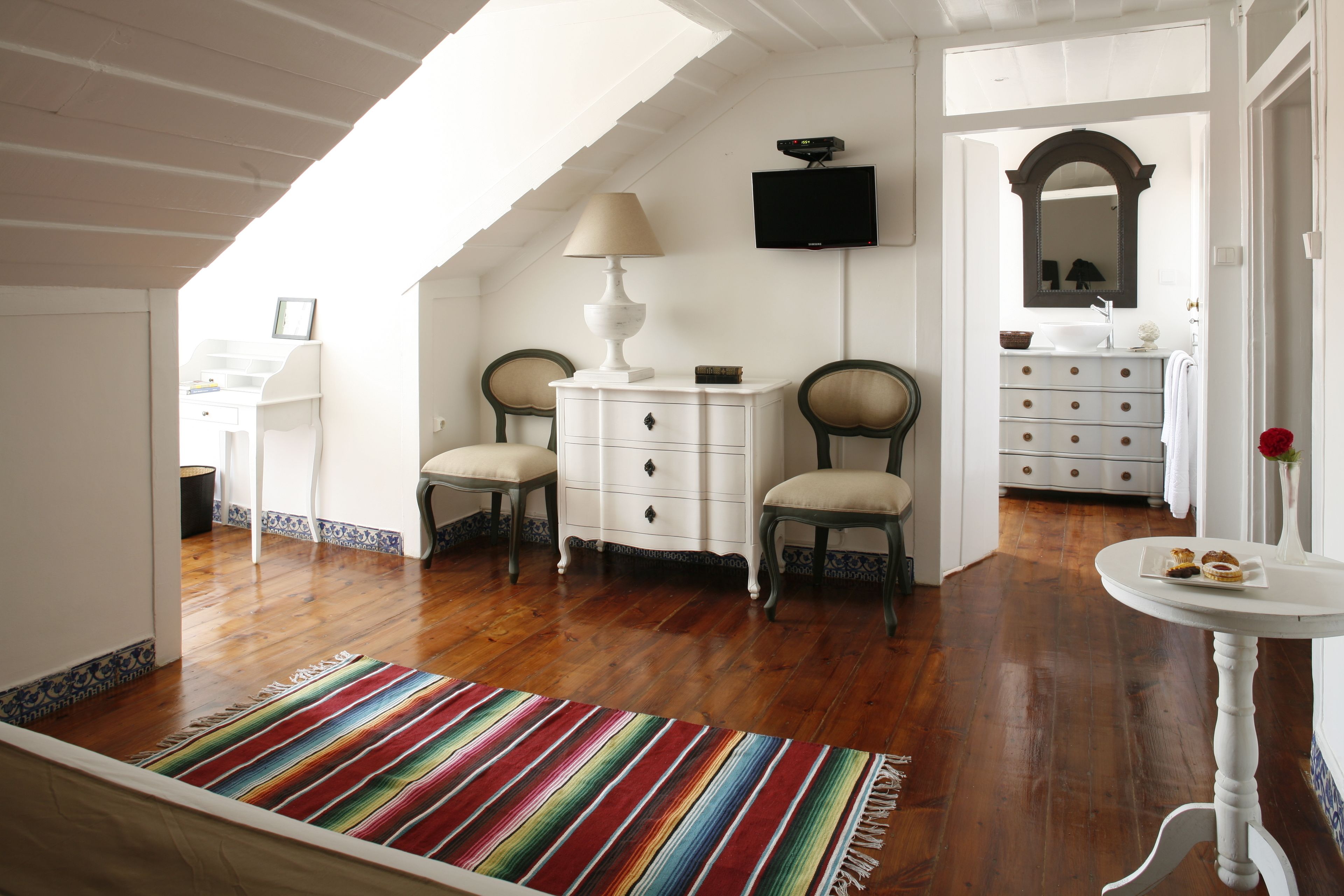 A room with a wooden floor, a colorful striped rug, a white dresser, two chairs, a small table with a lamp, and a television on the wall.