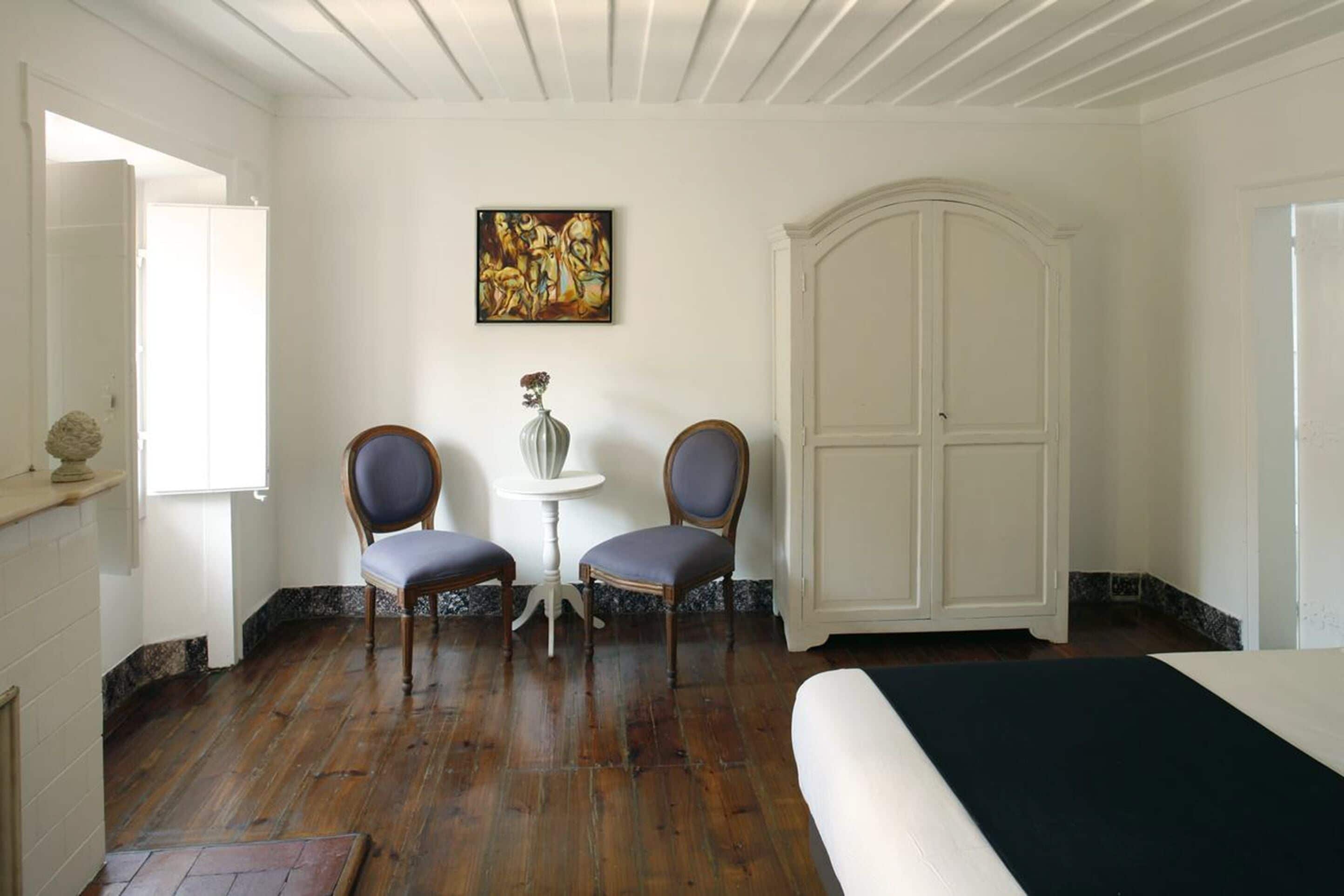 A room with wooden flooring, a bed, two chairs, a small table with a vase, a painting on the wall, and a large white cabinet.