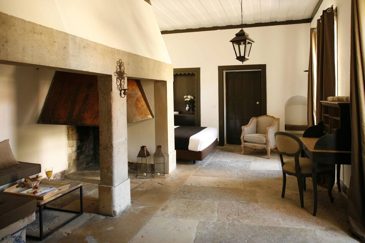 A cozy living room with a fireplace, a sofa, a dining table with chairs, and a bed in the background.