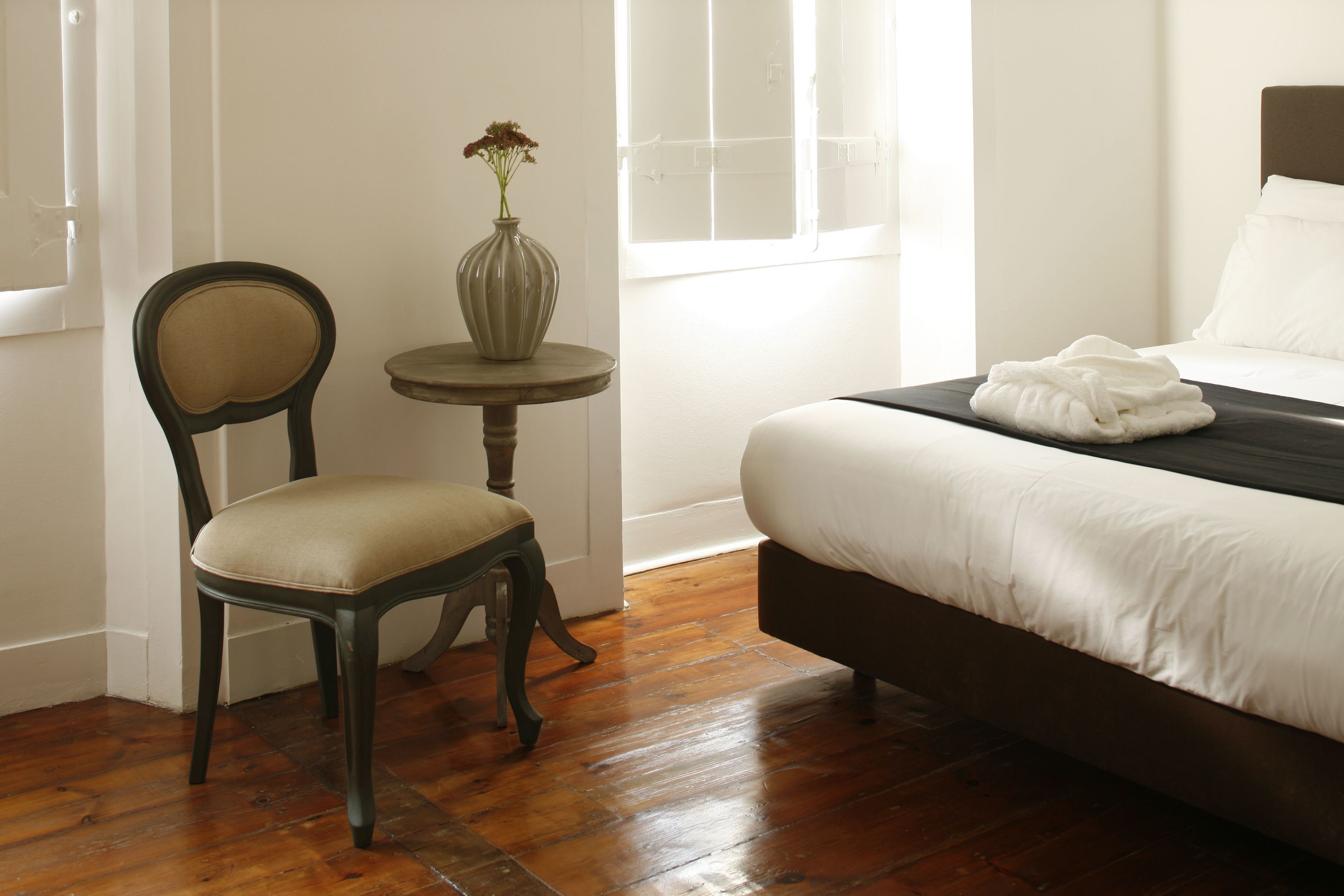 A hotel room with a bed, a chair, a small table with a vase, and a window.