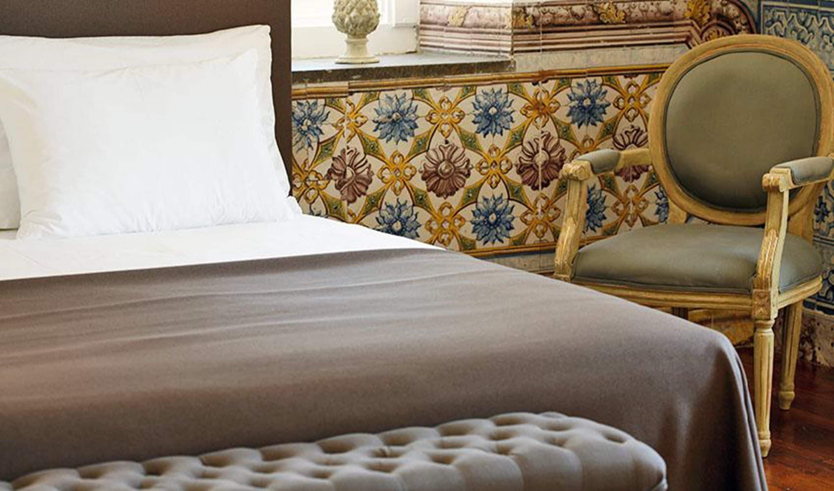 Una cama bien hecha con cubrecolcha marrón, una silla tapizada de verde y una pared de azulejos estampados.