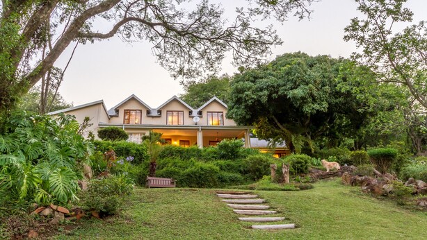 A two-story house with a covered porch, surrounded by lush greenery and a stone pathway leading to the entrance. - Porcupine Ridge Guest House (Sabie)