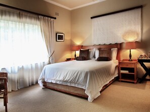 A neatly made bed with white linens, two bedside tables with lamps, a wooden nightstand, a wicker chair, and a large window with sheer curtains. - Porcupine Ridge Guest House (Sabie)