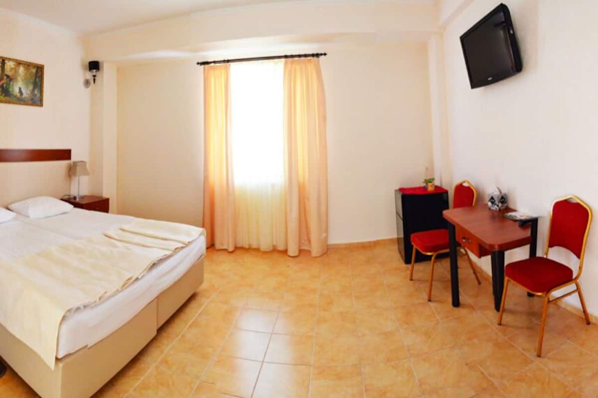 A hotel room with a bed, a desk with two chairs, a TV, and a window with curtains.