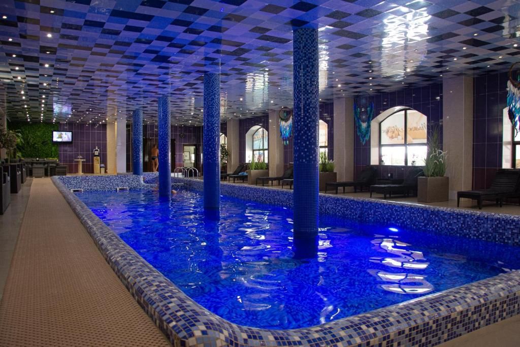 An indoor swimming pool with a checkered ceiling, tiled walls, and a seating area with tables and chairs.