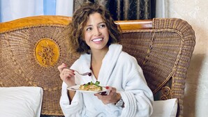 Una persona sentada en una cama dentro de una habitación de hotel, con una bata blanca y sosteniendo un plato de comida.