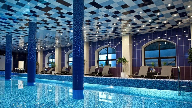 Una piscina interior con azulejos de techo a cuadros, suelo de mosaico azul y sillones de descanso junto a la piscina.