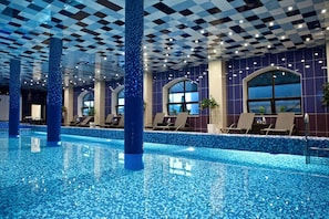 An indoor swimming pool with checkered ceiling tiles, blue mosaic pool floor, and lounge chairs along the poolside.
