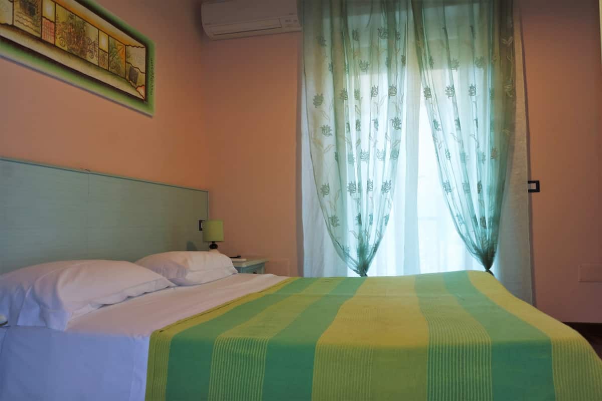 Una habitación de hotel con cama, mesita de noche, lámpara y ventana con cortinas traslúcidas.