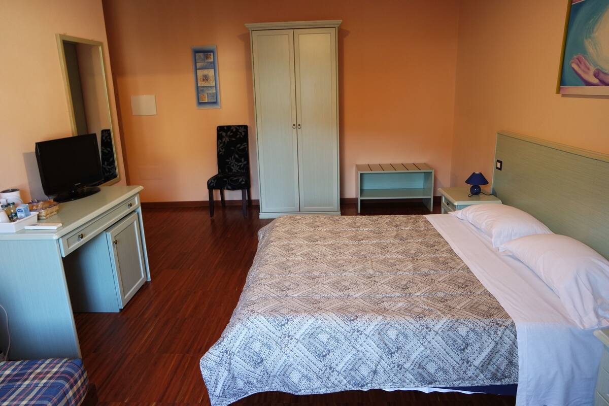Un dormitorio con una cama, un televisor sobre un escritorio, una silla, un armario y una mesita de noche con una lámpara.