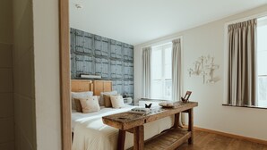 Un dormitorio con cabecera de madera, una banca de madera, cama con ropa de cama blanca y una ventana con cortinas.