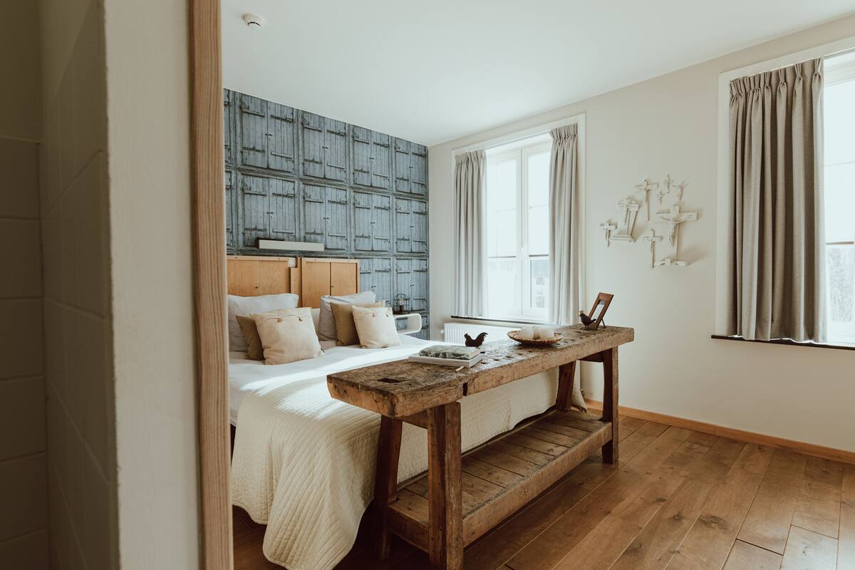 Un dormitorio con cabecera de madera, una banca de madera, cama con ropa de cama blanca y una ventana con cortinas.