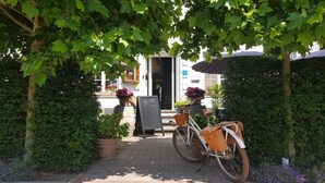 Una bicicleta estacionada bajo un árbol, un cartel con la palabra “WELKOM” escrita y la entrada de un edificio con una puerta.