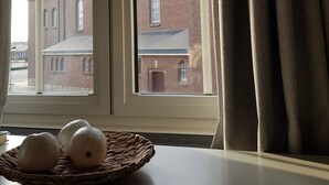 Una ventana con vistas a un edificio de ladrillo, una mesa blanca con una cesta de pan y una cortina entreabierta.