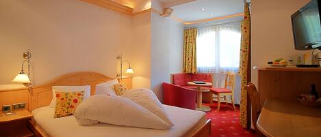 Habitación de hotel con cama doble, dos mesitas de noche con lámparas, una silla roja pequeña, una mesa redonda y un televisor sobre un mueble de madera.