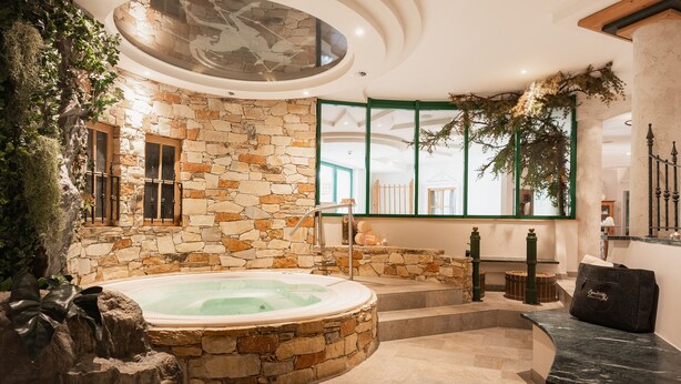 A stone-walled indoor pool area with a hot tub, steps leading to a balcony, and a view of the outdoors through large windows. - Vitalhotel Samnaunerhof  (Samnaun)
