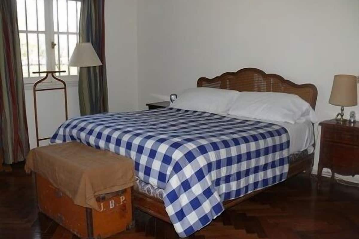 Un dormitorio con cama de madera, una mesita de noche, una lámpara, un ventanal con cortinas y un banco de madera.