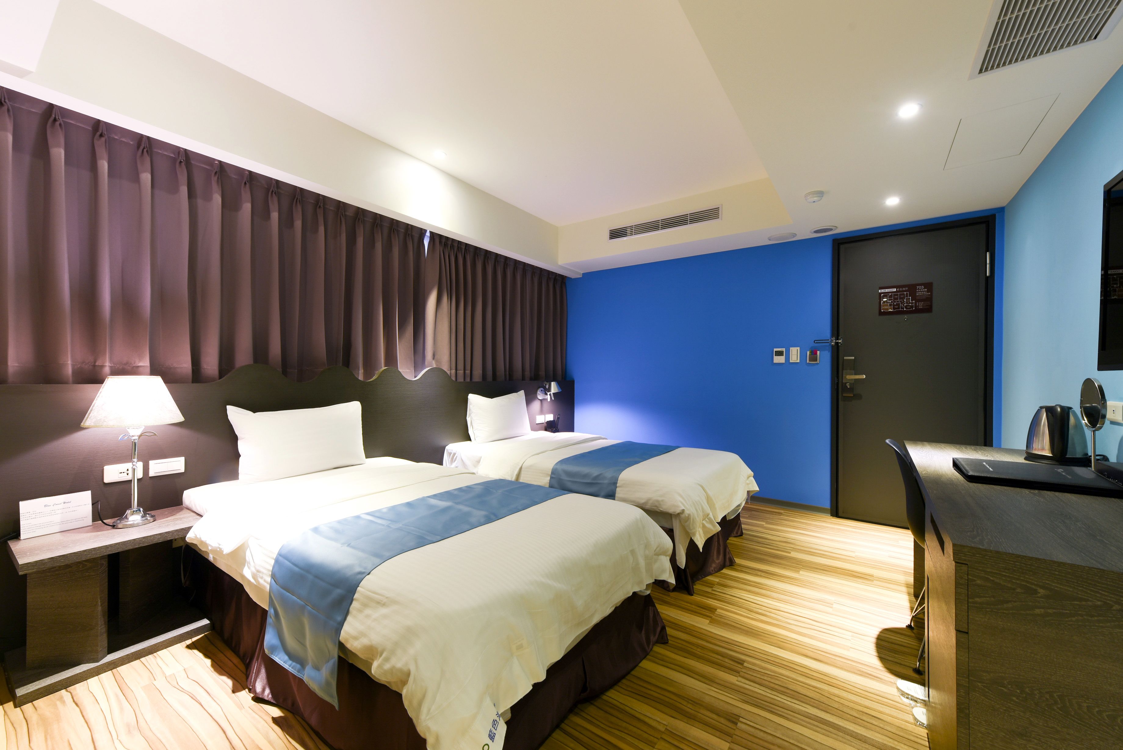 A hotel room with two beds, a desk, a lamp, and a door with a sign.