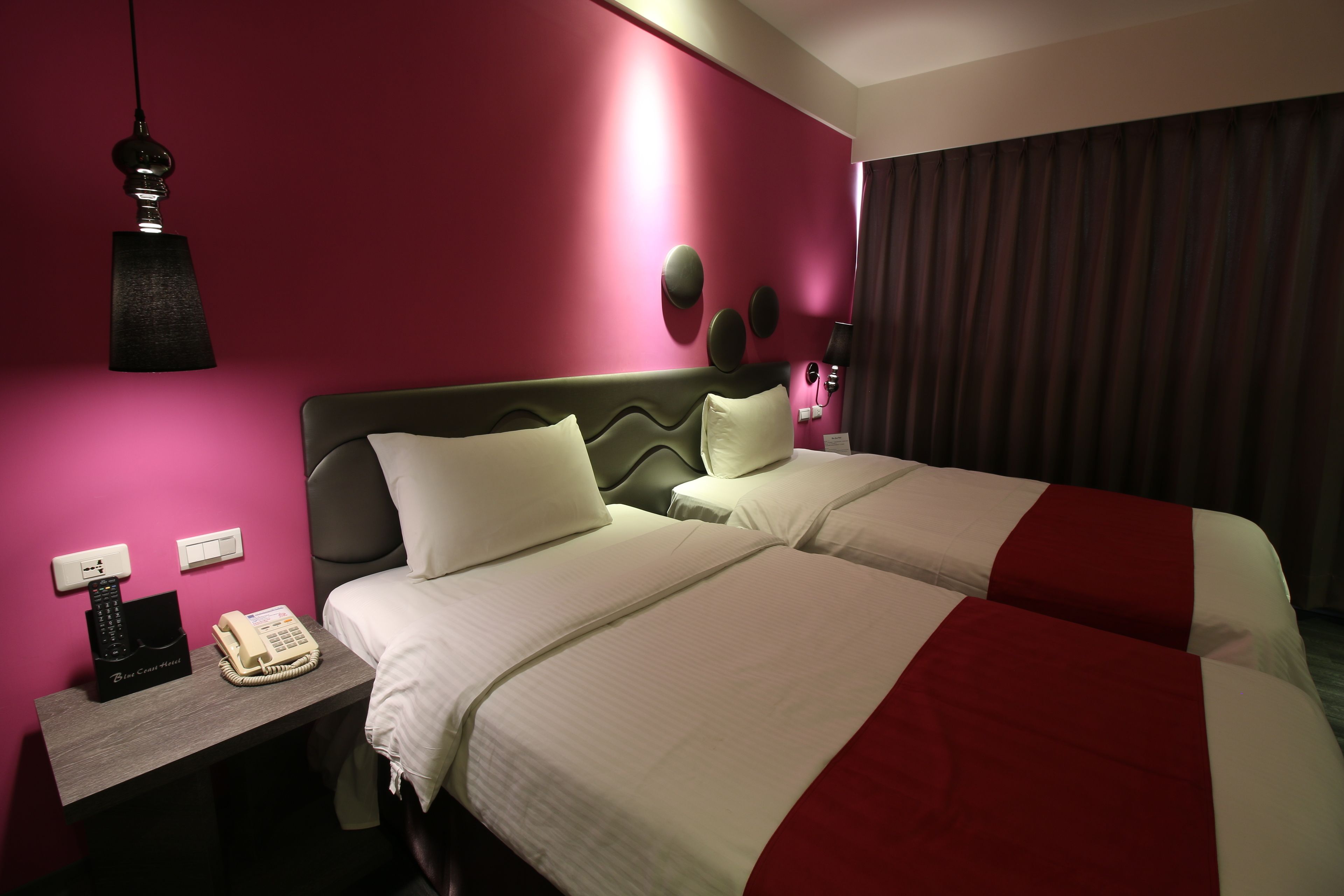 A hotel room with two beds, a bedside table with a telephone, and a wall-mounted lamp.