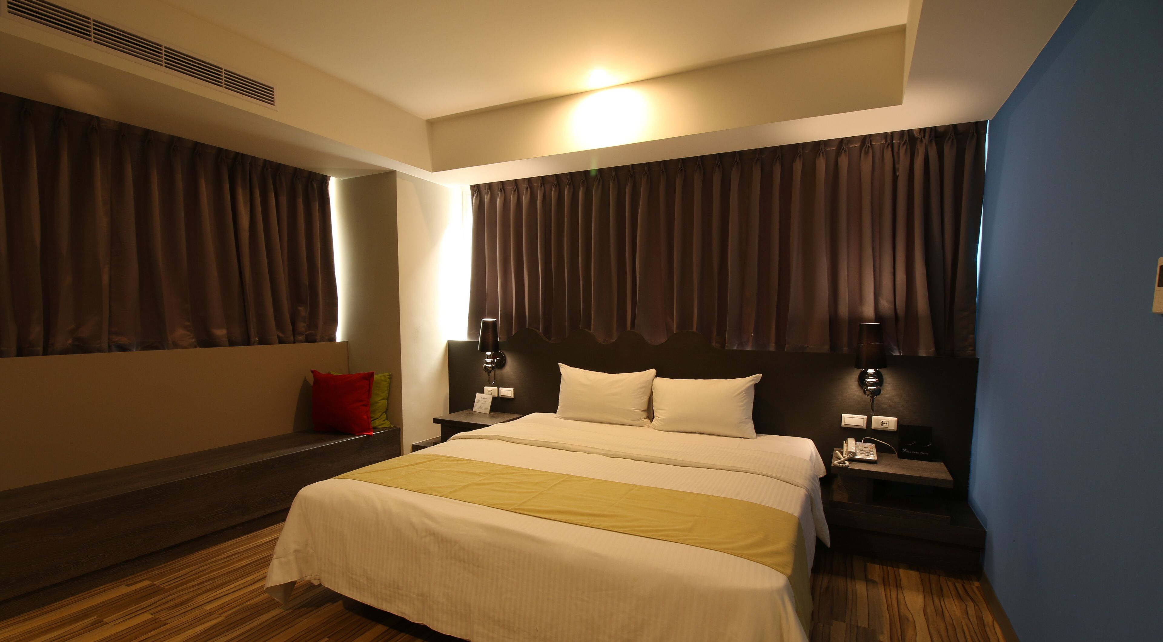 A hotel room with a large bed, two bedside tables, a nightstand, a wall-mounted lamp, and a window with curtains.