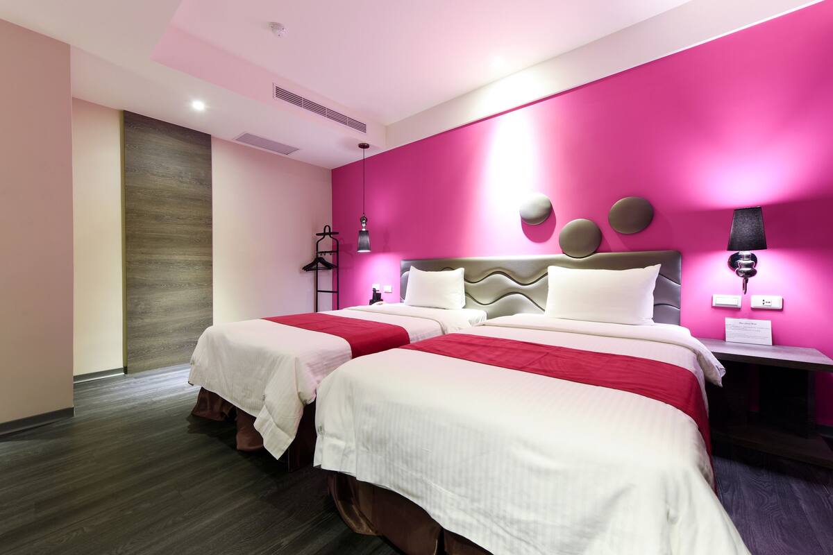 Ein Hotelzimmer mit zwei Betten, Holzboden, einer Wand in Rosa und einem Nachttisch mit Lampe.