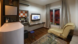 Ein Wohnzimmer mit Fernseher, einer Couch, einem Glastisch, einer Küchenzeile inklusive Kühlschrank und einem Fenster mit Vorhängen.