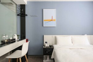 Een hotelkamer met een bed, een bureau met stoel, een nachtkastje met telefoon en lamp, een lijst op de muur, en een raam met uitzicht op zee.