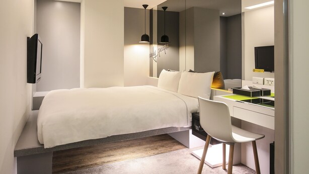 A modern bedroom with a bed, a desk, and a chair. There is a mirror above the desk and a TV on the wall.
