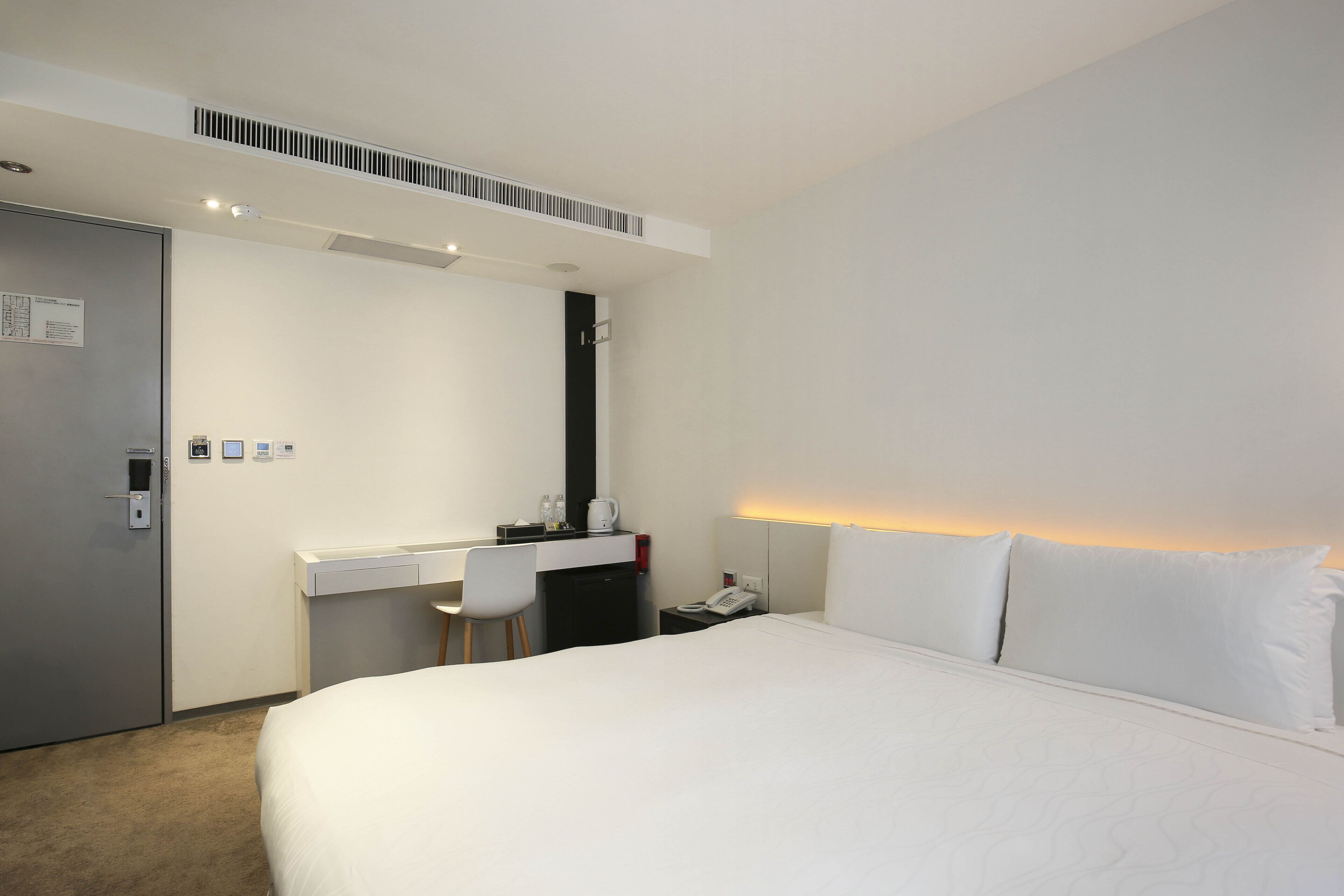 A hotel room with a bed, a desk, a chair, a nightstand, and a door.