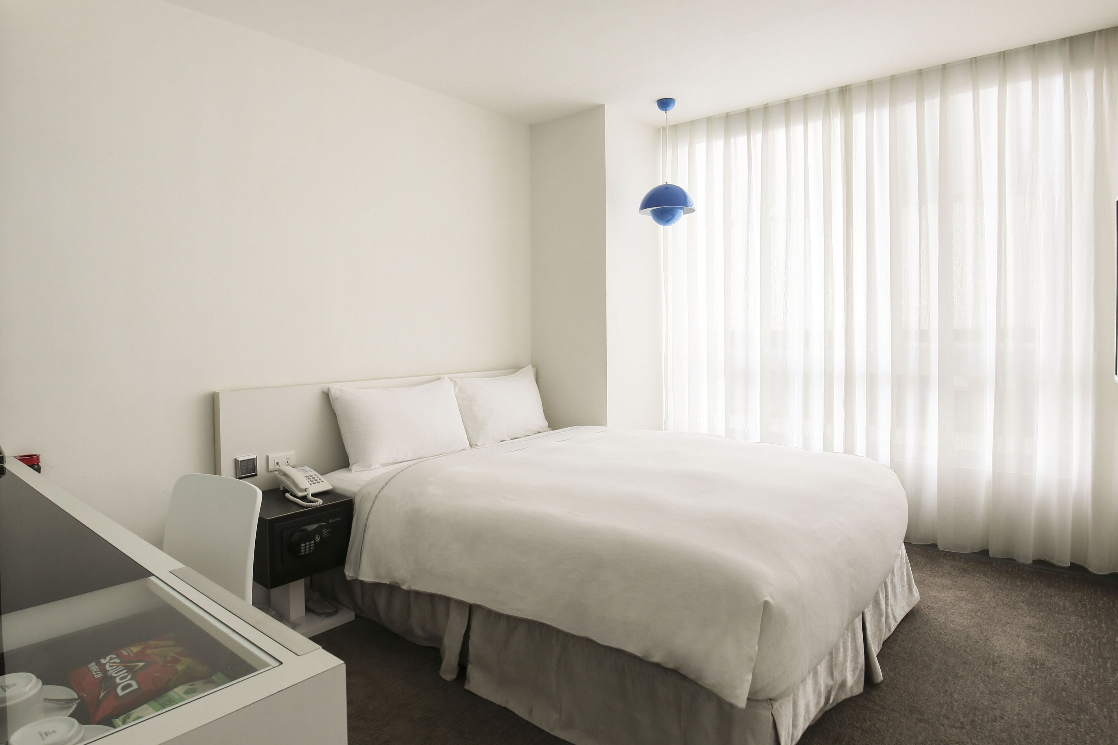 Une chambre d’hôtel avec un grand lit, une table de chevet avec un téléphone et une lampe, un bureau avec un ordinateur et une fenêtre avec des rideaux légers.