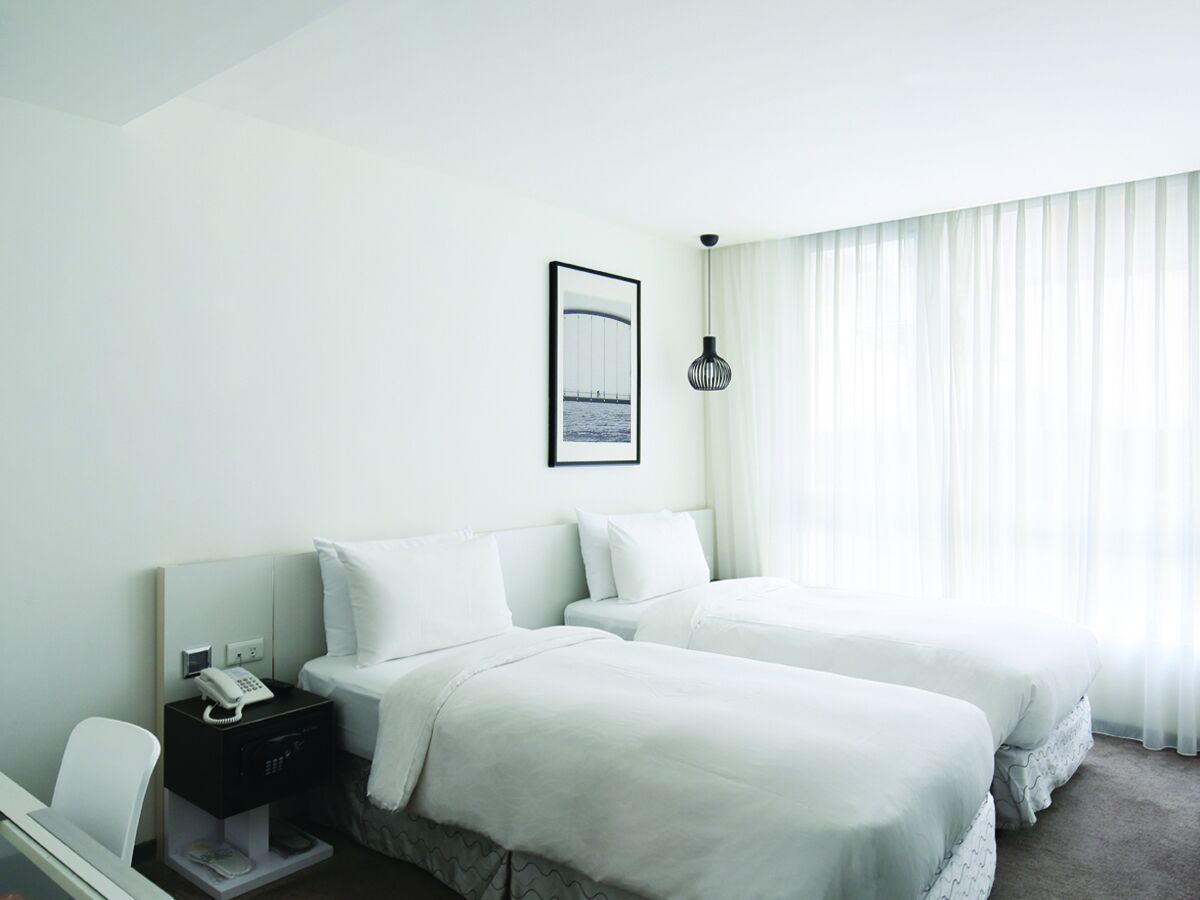 A hotel room with two beds, a bedside table with a phone, a framed picture on the wall, and sheer curtains on the window.