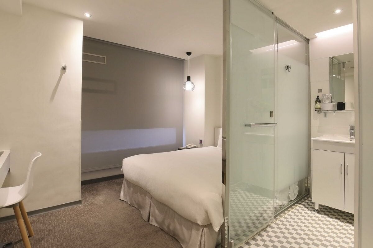Una habitación de hotel moderna con cama, ducha con cerramiento de vidrio, una silla blanca y un escritorio pequeño.