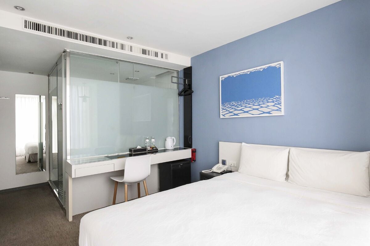 A modern hotel room with a large bed, a desk with a chair, a glass shower enclosure, and a painting on the wall.