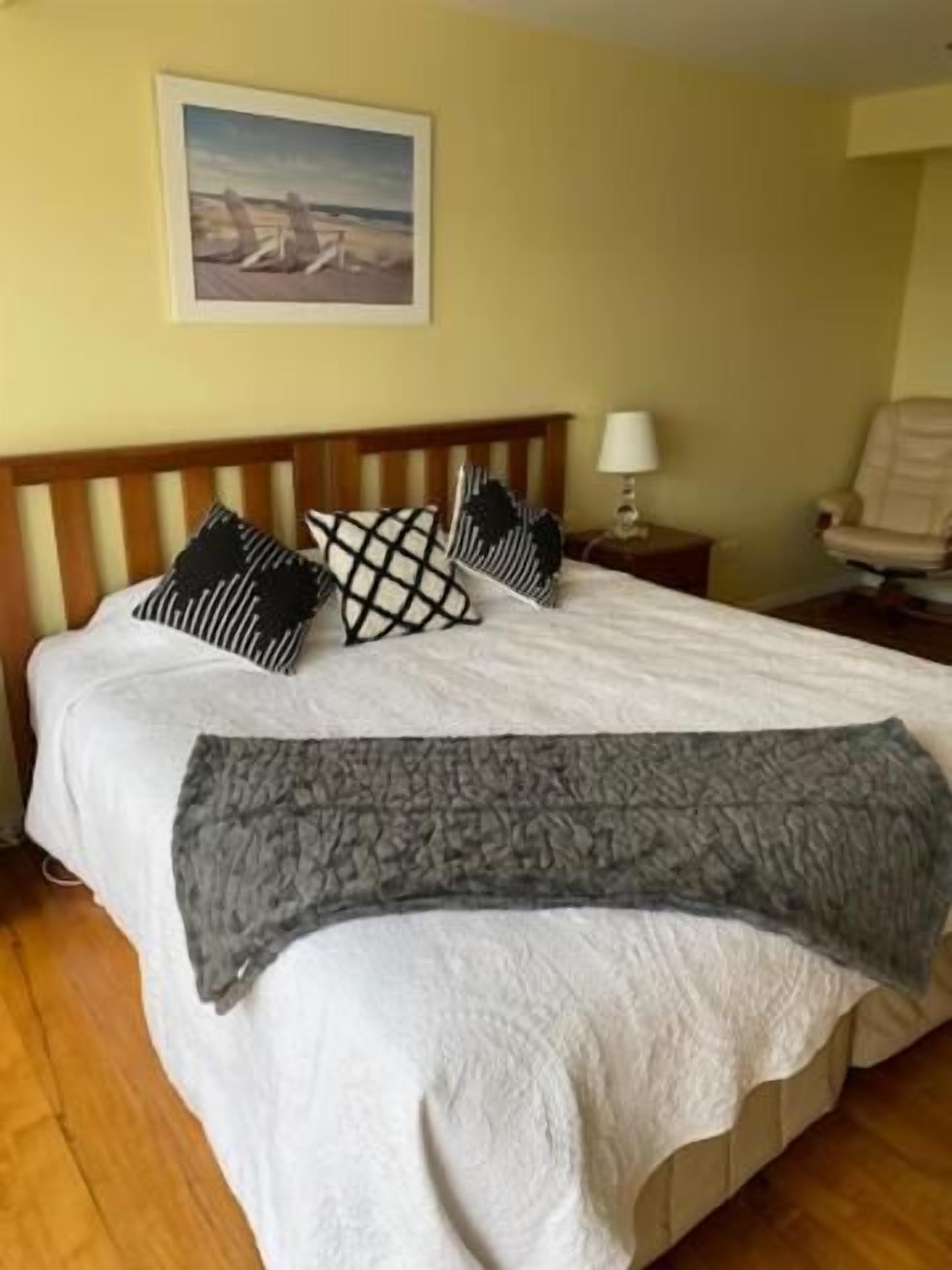 A neatly made bed with a white duvet, a grey throw blanket, and patterned pillows. There is a wooden headboard, a bedside lamp, and a chair in the room.
