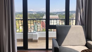 A room with a large window, a grey armchair, and a view of a cityscape with buildings and greenery. - Thi Thao Gardenia Hotel (Xuan Huong - Da Lat)