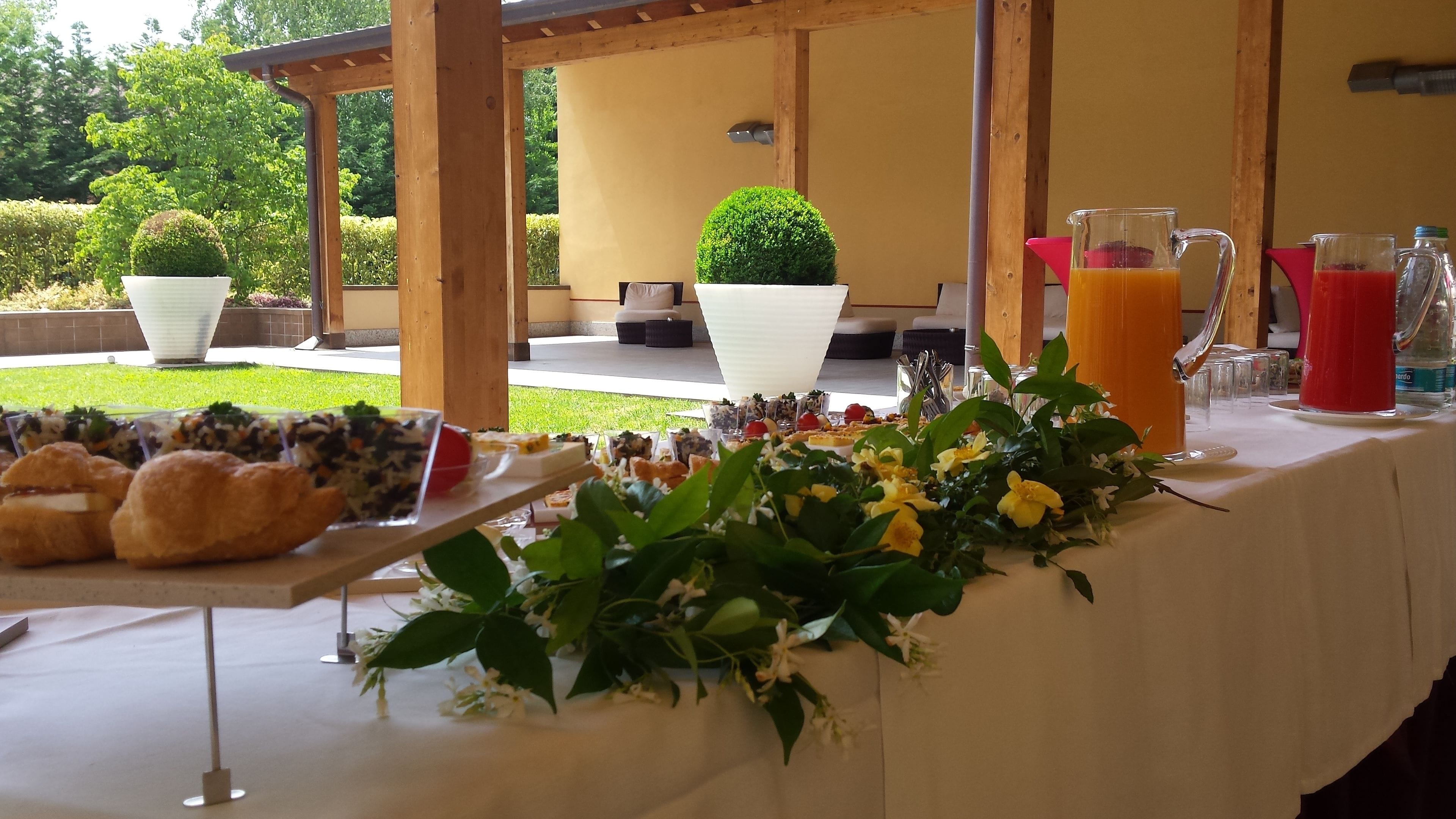A breakfast buffet with various food items and drinks, set up outdoors with potted plants and a wooden pergola.