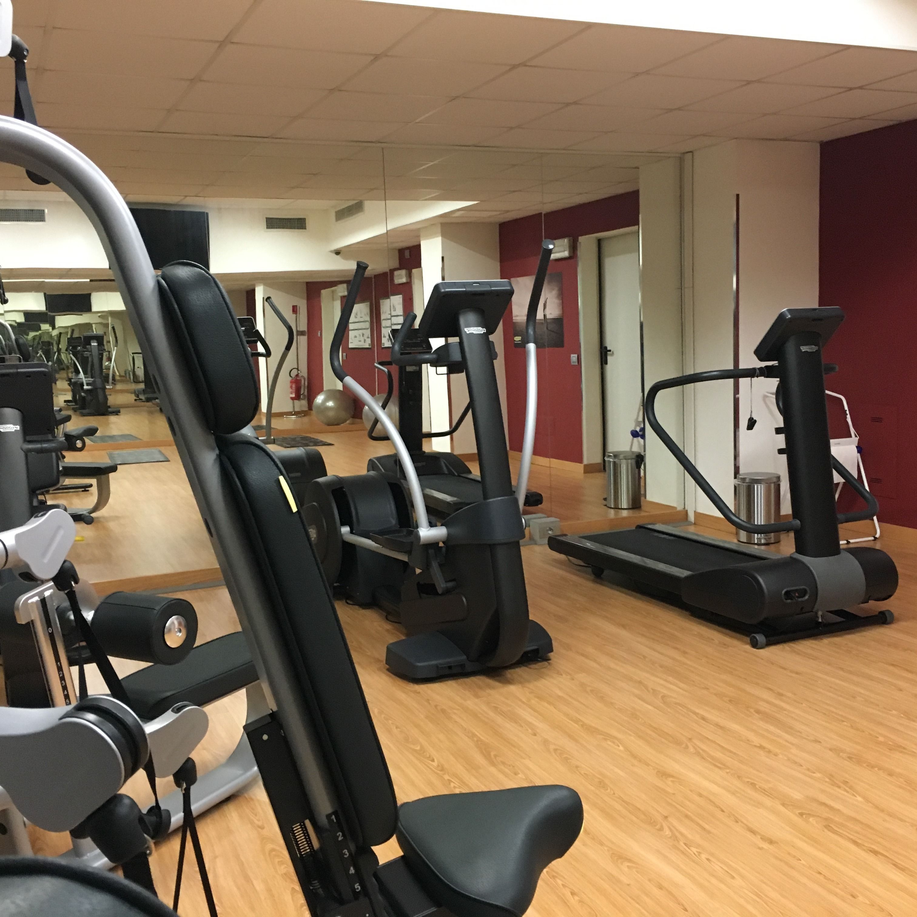 A gym with various exercise machines, including treadmills and elliptical trainers, arranged in a spacious room with wooden flooring and mirrored walls.