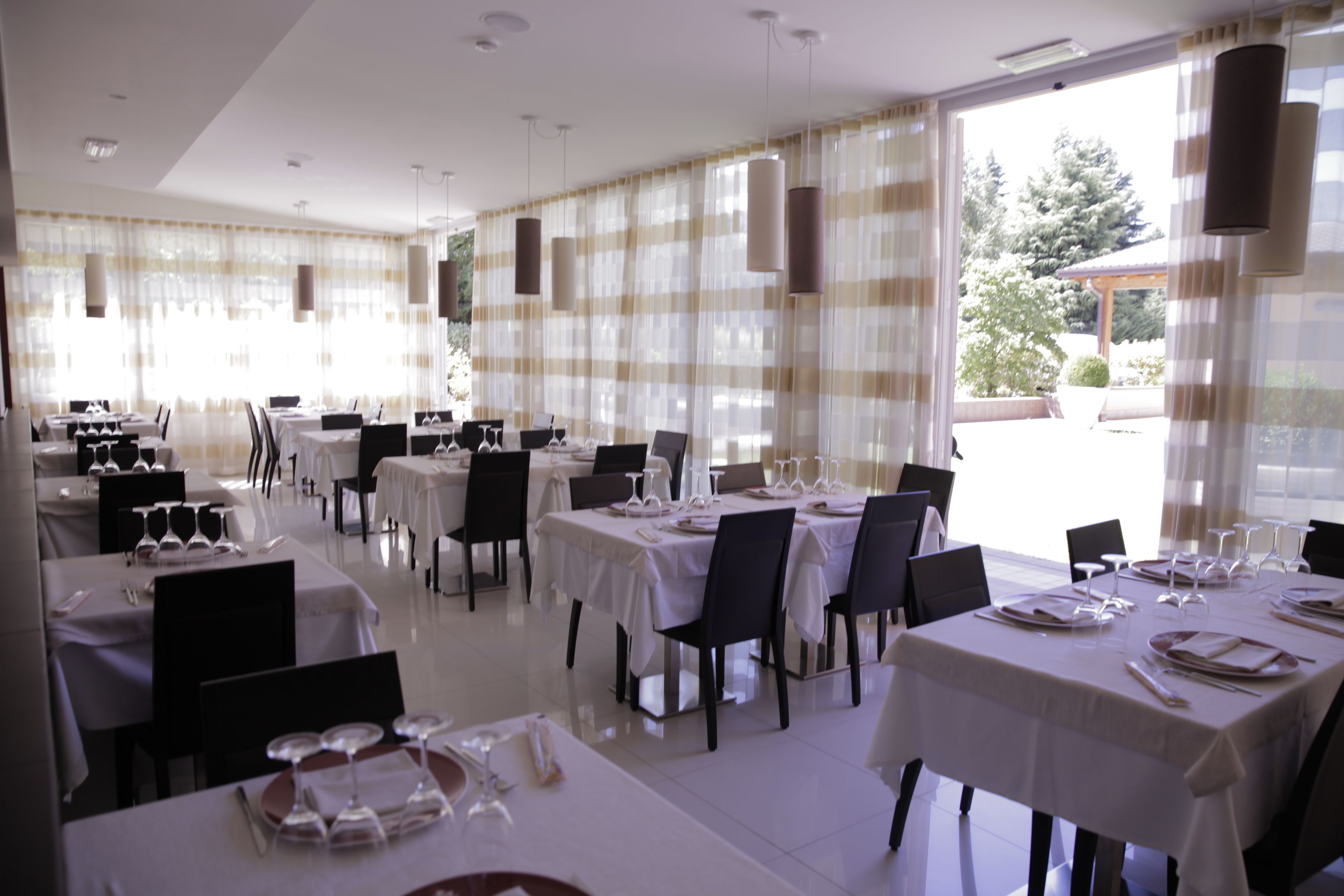 A dining area with tables set for service, large windows with curtains, and a view of the outdoors.